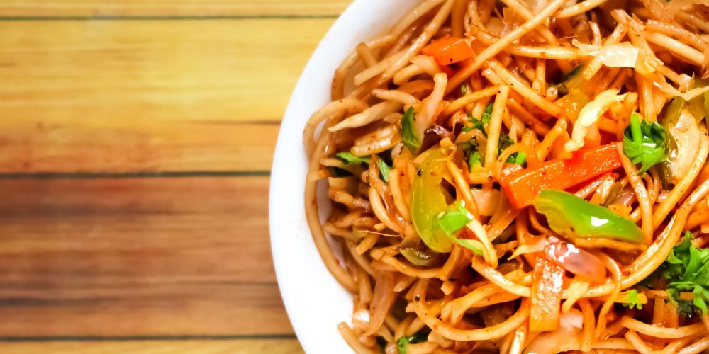 A vibrant bowl of spicy Indian noodles garnished with vegetables on a wooden table.