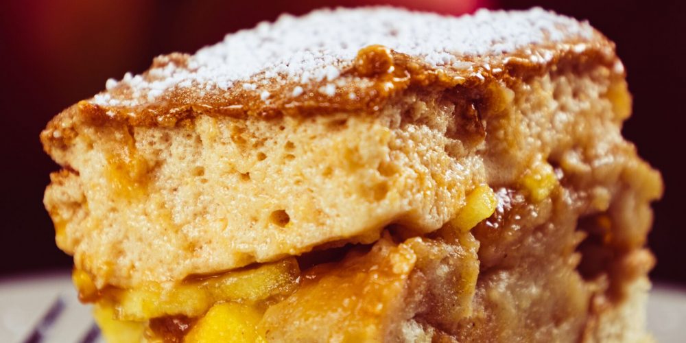 A square serving of apple cobbler with a soft golden baked topping over cinnamon-spiced apples, dusted with powdered sugar.